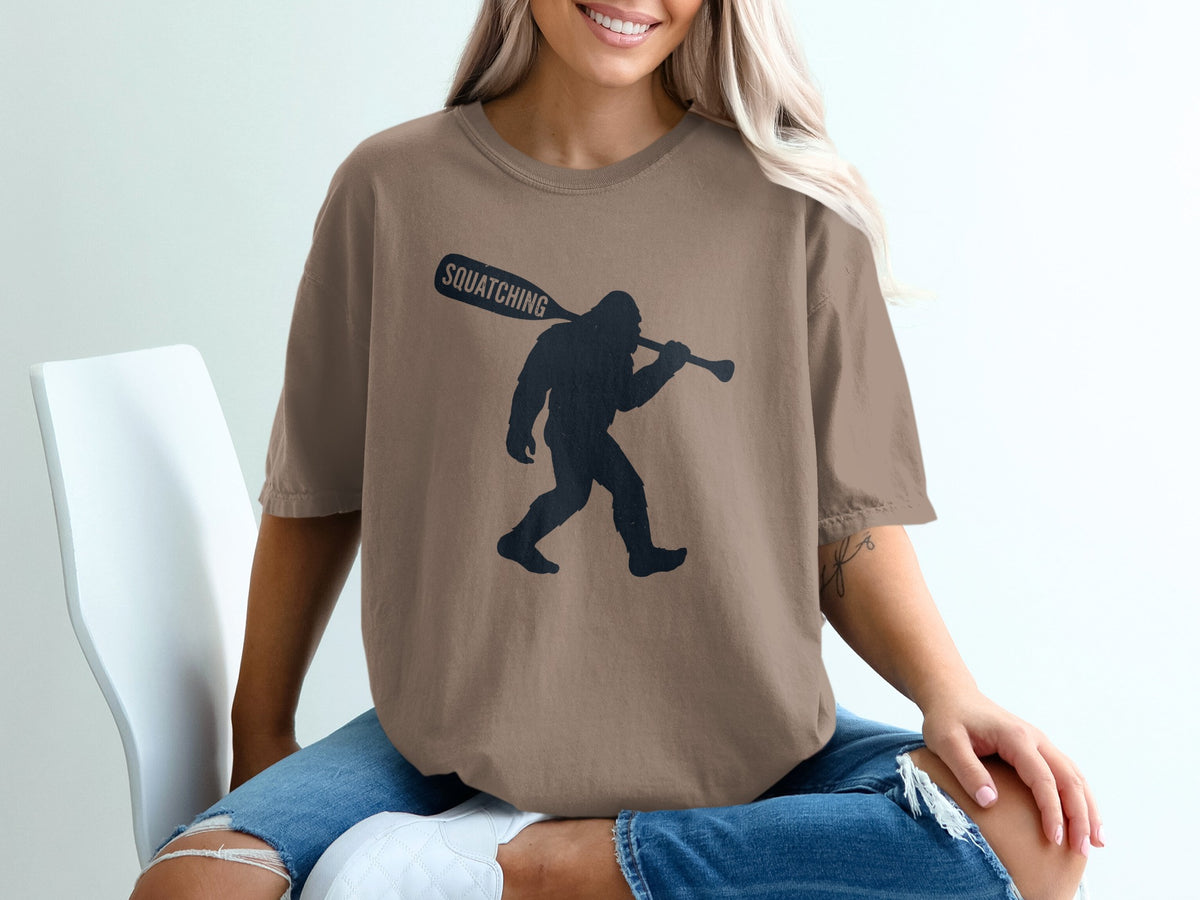 Brown t-shirt with black Bigfoot silhouette carrying "Squatching" paddle.