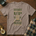 A tan t-shirt with olive green text reading Hurry Up and Wait surrounding an hourglass graphic.