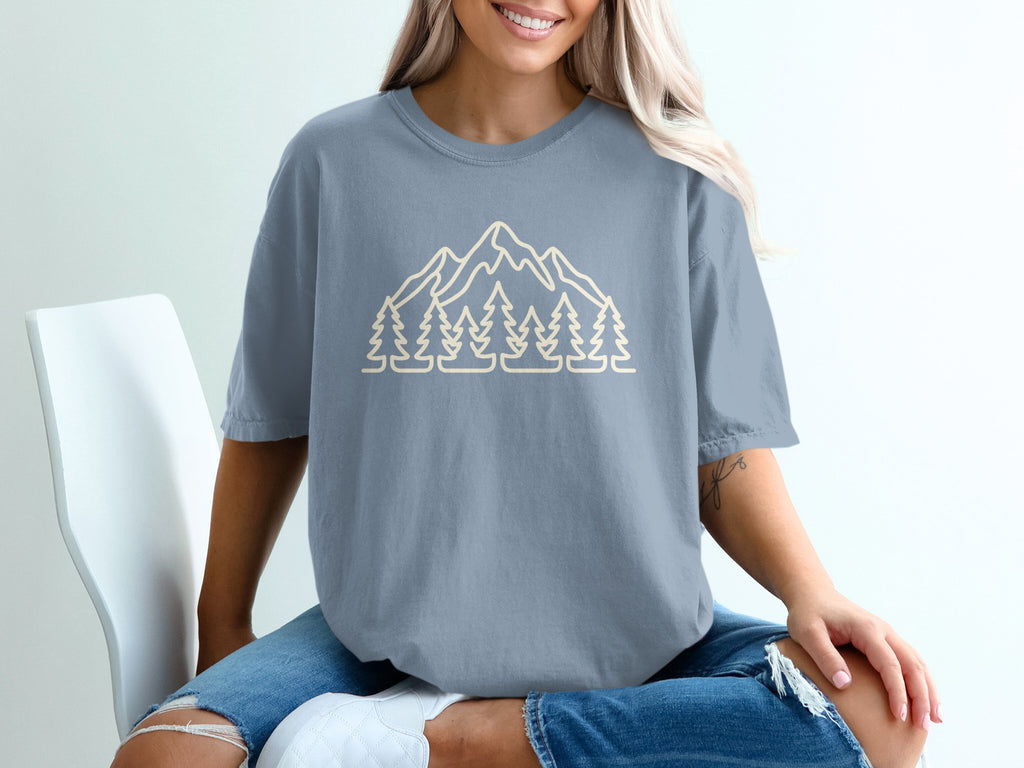 Light beige outline graphic of mountains and pine trees on a slate blue t-shirt.