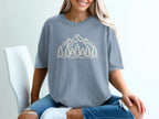 Light beige outline graphic of mountains and pine trees on a slate blue t-shirt.