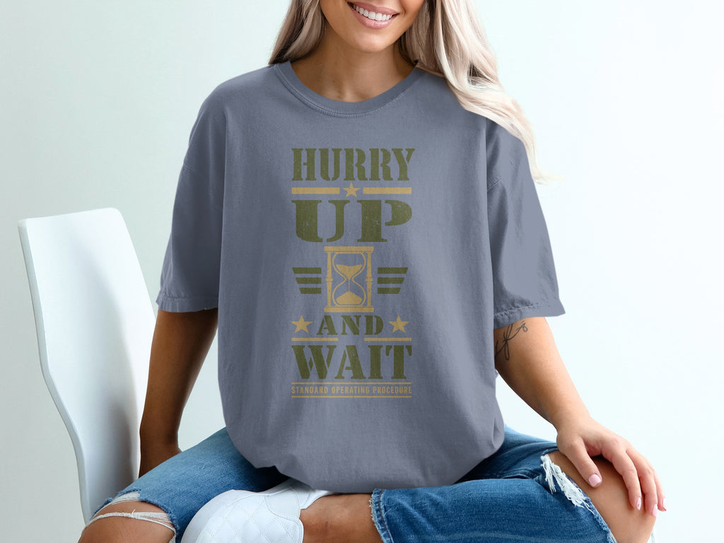 A woman wearing a gray t-shirt with olive green text that reads Hurry Up And Wait.