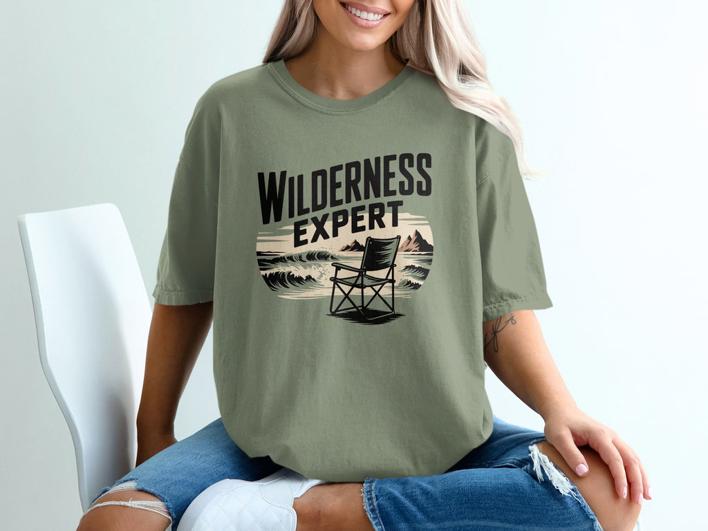 Olive green t-shirt with "WILDERNESS EXPERT" text and beach scene graphic.