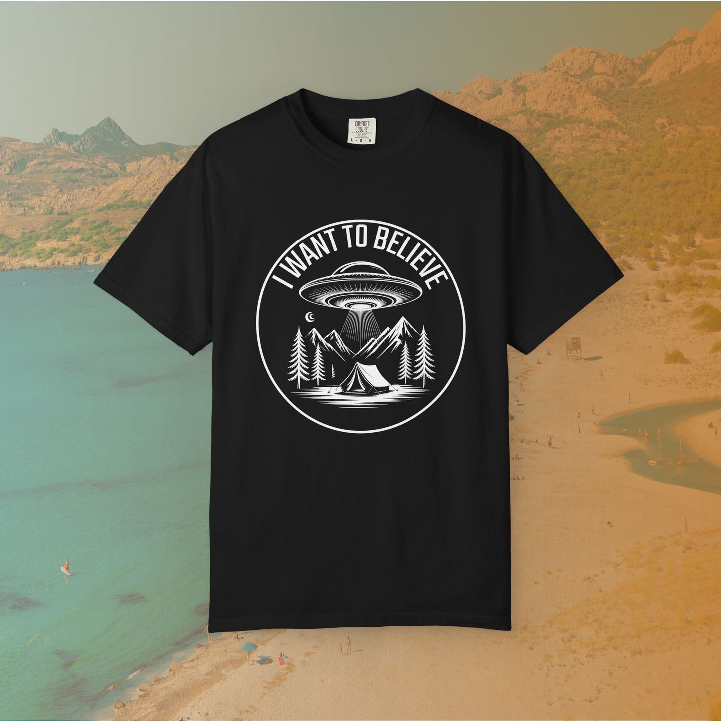  t-shirt A round, white graphic on a dark background featuring a classic flying saucer UFO abducting a tent at a campsite surrounded by mountains and pine trees. The surrounding text reads "I WANT TO BELIEVE.