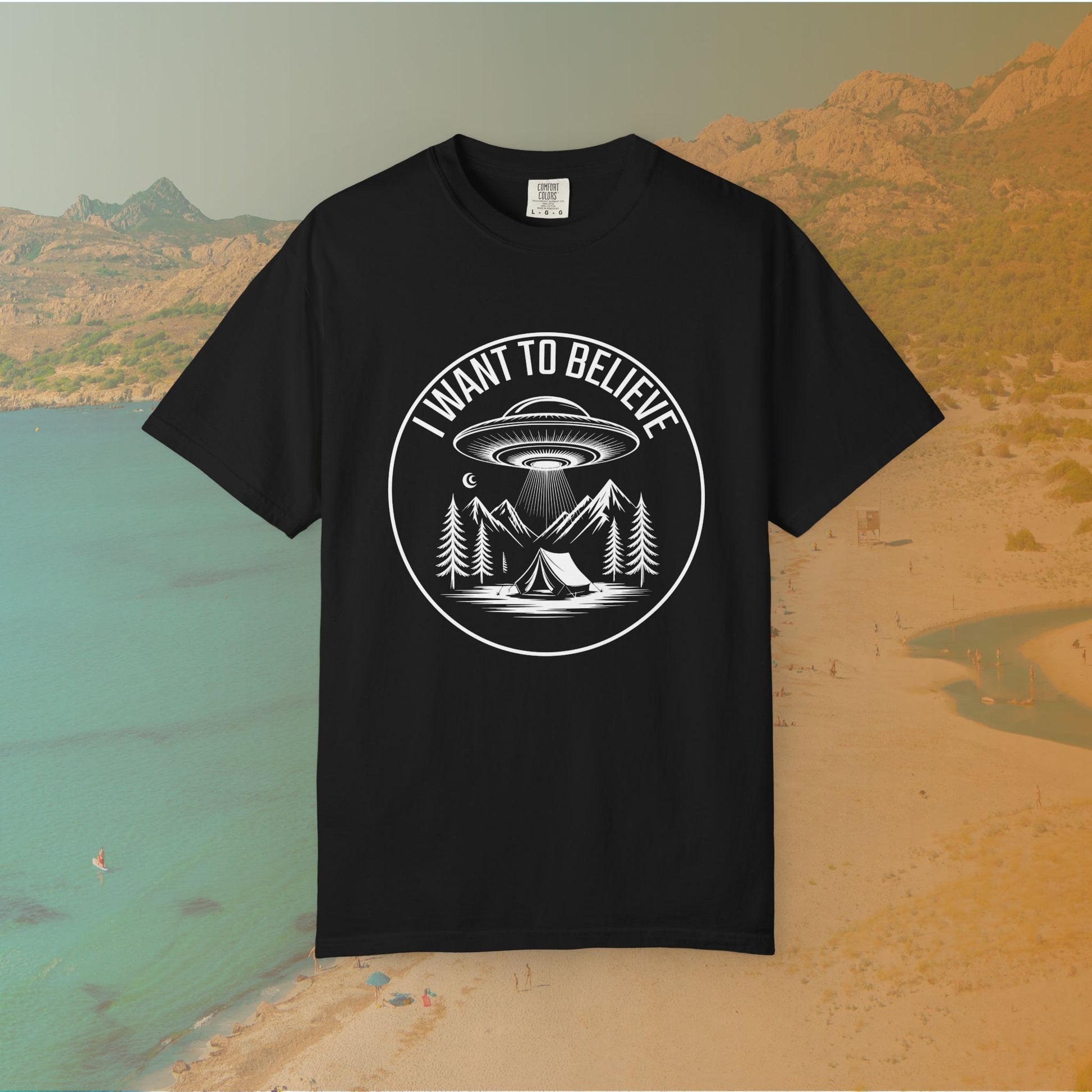  t-shirt A round, white graphic on a dark background featuring a classic flying saucer UFO abducting a tent at a campsite surrounded by mountains and pine trees. The surrounding text reads "I WANT TO BELIEVE.