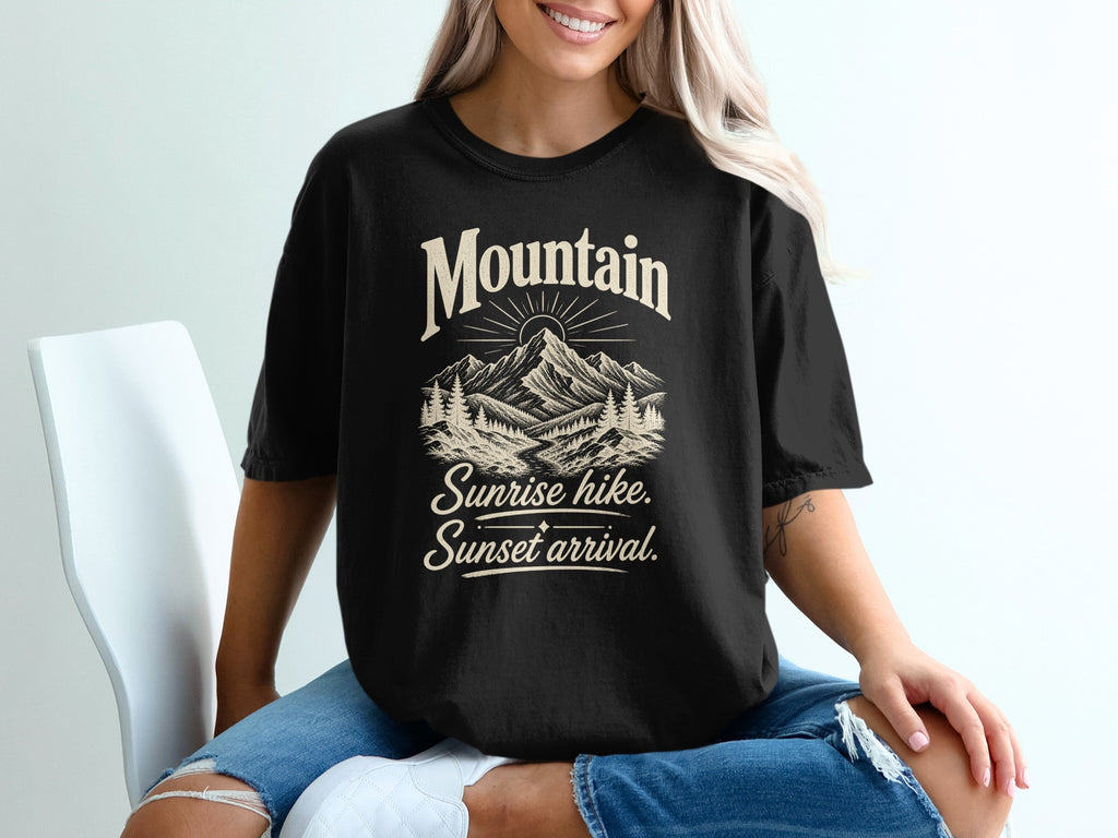 A blonde woman wearing a black t-shirt printed with a mountain graphic and the text Mountain Sunrise hike Sunset arrival.