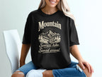 A blonde woman wearing a black t-shirt printed with a mountain graphic and the text Mountain Sunrise hike Sunset arrival.