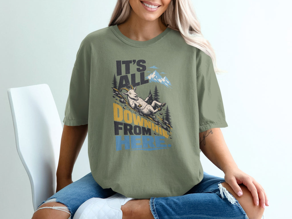 A woman wearing an olive-green t-shirt printed with a mountain scene, goat, and text reading It's All Downhill From Here.