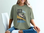 A woman wearing an olive-green t-shirt printed with a mountain scene, goat, and text reading It's All Downhill From Here.