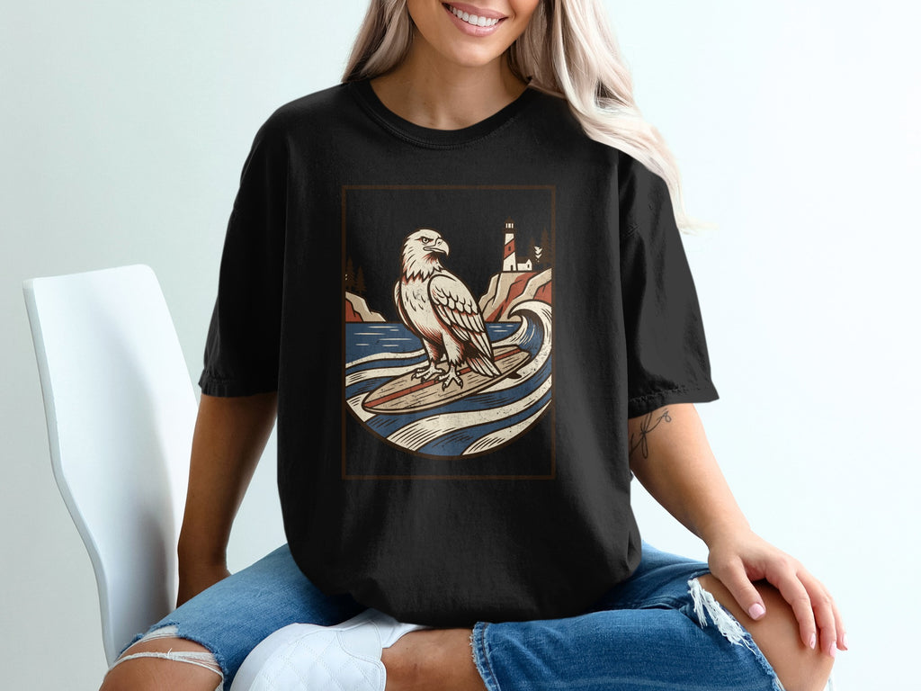 Black t-shirt with a graphic of an eagle surfing on a wave near a lighthouse.
