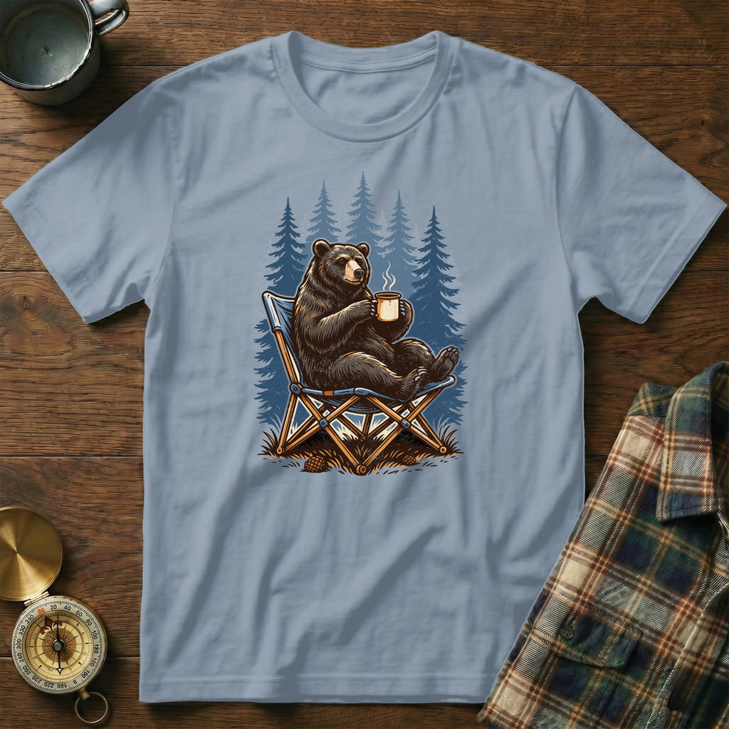 Bear in camping chair holding a mug in front of pine trees.