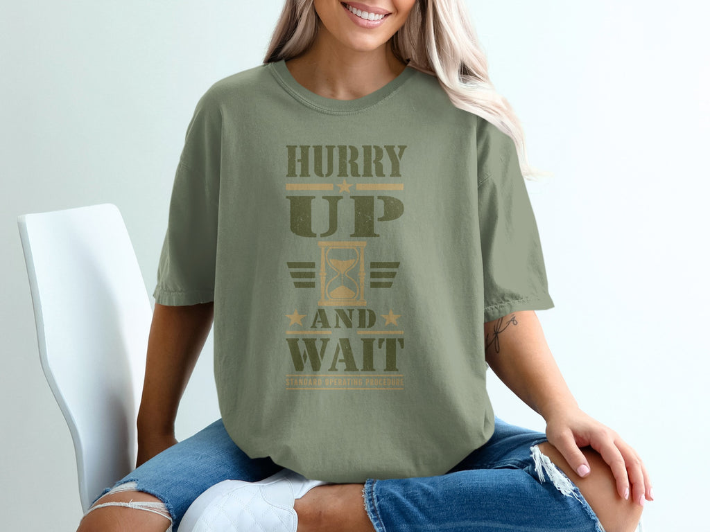 A woman sits wearing an olive green t-shirt with "Hurry Up and Wait" printed in distressed text.