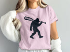 Bigfoot silhouette on a pink t-shirt carrying a paddle with "Squatching" text.