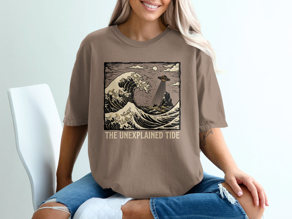 T-shirt graphic featuring a wave, UFO, and text "The Unexplained Tide".