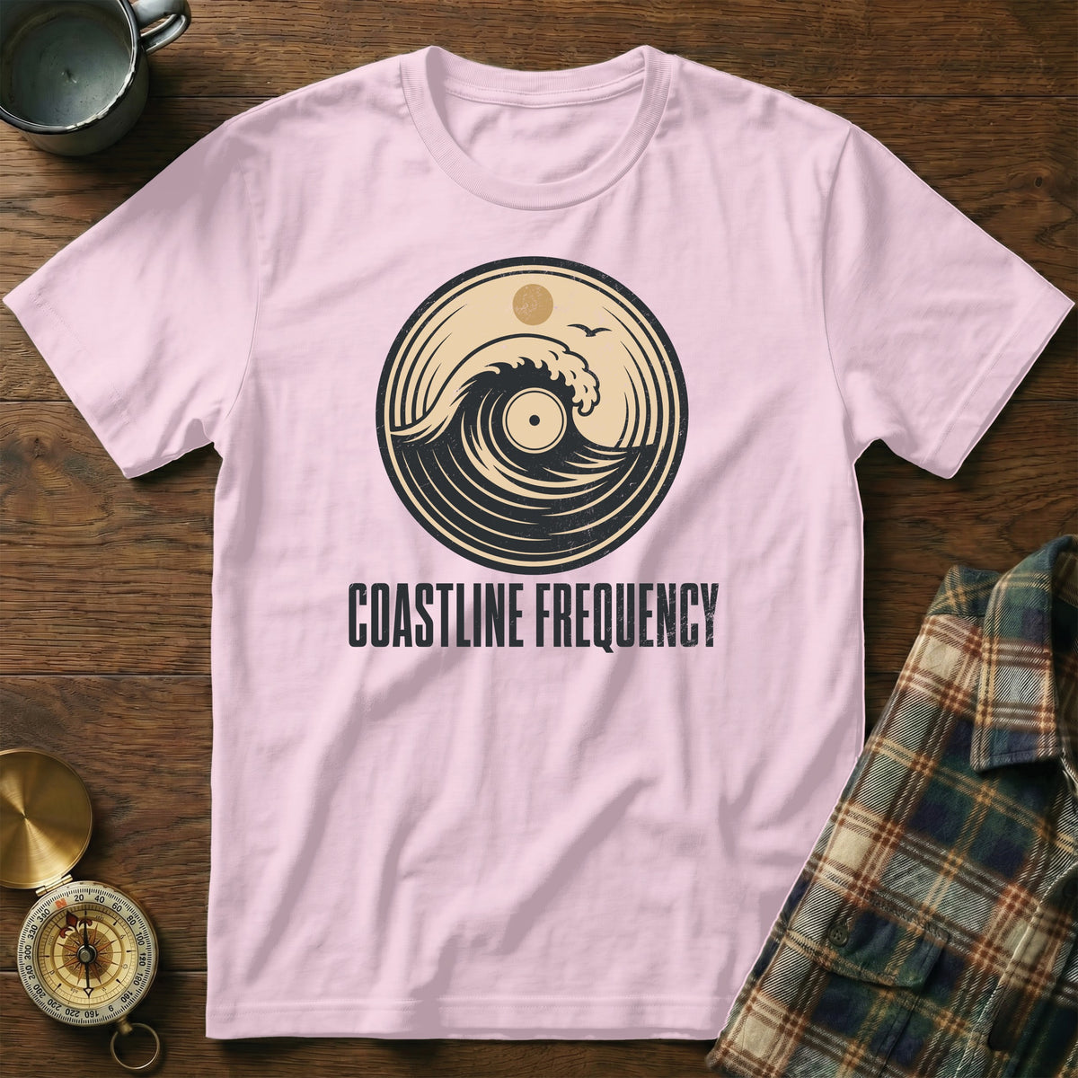 Pink t-shirt with a circular logo featuring a wave, sun, bird, and the text COASTLINE FREQUENCY.