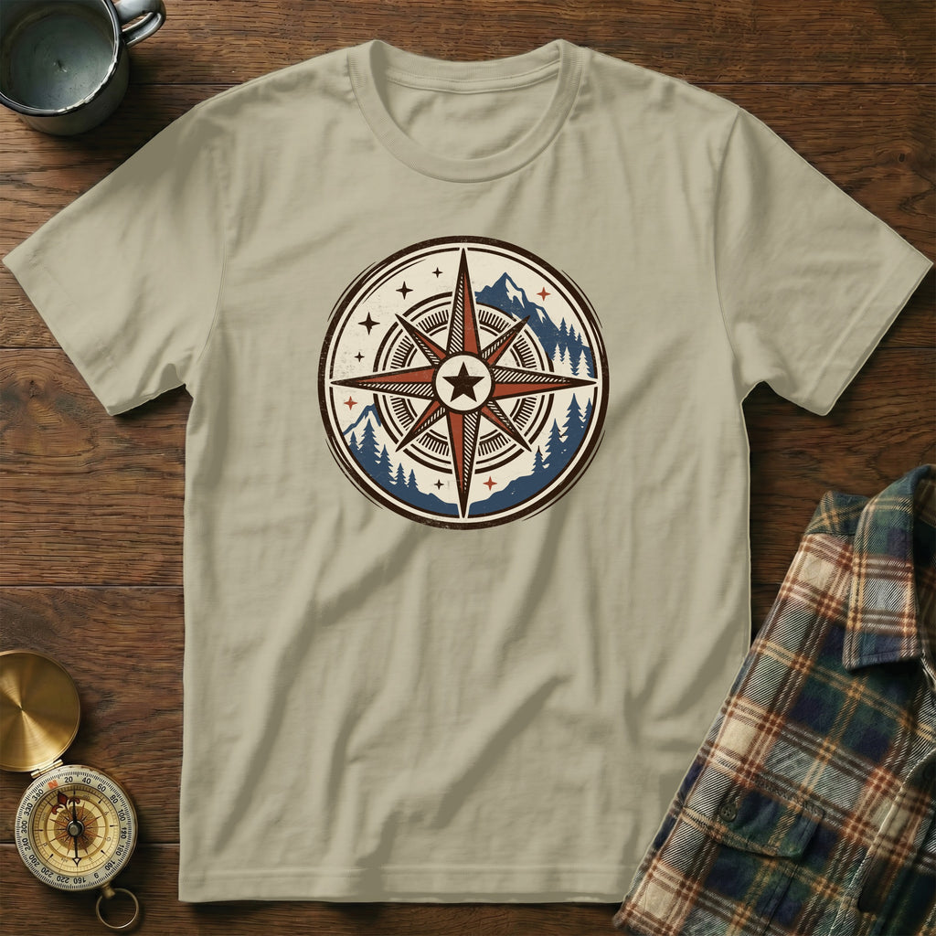 T-shirt with circular compass logo, mountains, and pine trees, laid on wooden surface.