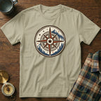 T-shirt with circular compass logo, mountains, and pine trees, laid on wooden surface.