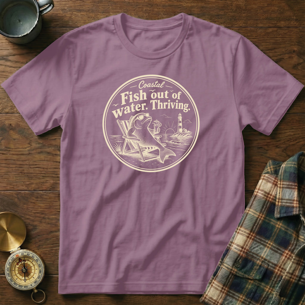 A purple t-shirt featuring a circular graphic with a fish relaxing in a beach chair and text reading Fish out of water. Thriving.