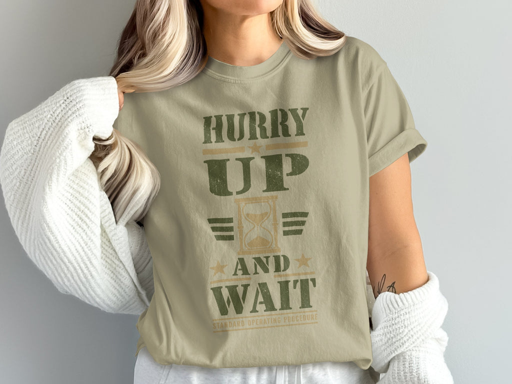 A person wearing a sage green t-shirt with distressed green text reading Hurry Up And Wait.
