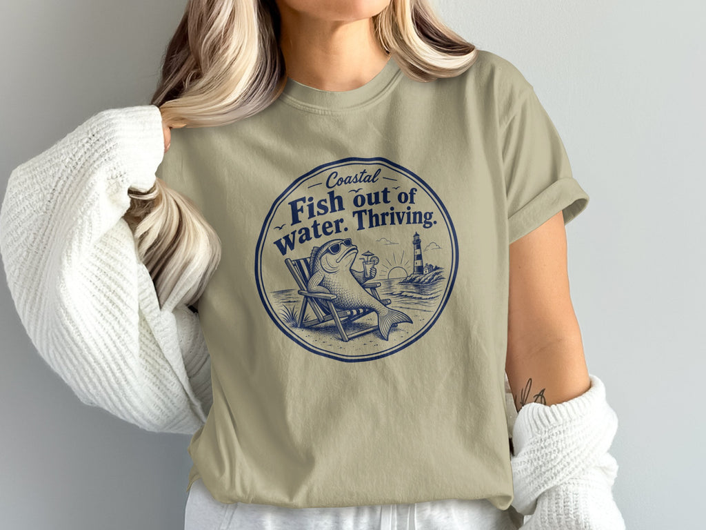 A woman wears a beige t-shirt featuring a blue graphic of a fish lounging in a beach chair with a drink.