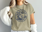 A woman wears a beige t-shirt featuring a blue graphic of a fish lounging in a beach chair with a drink.