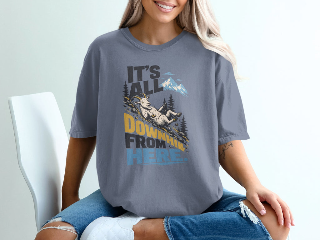 A blonde woman wears a grey t-shirt featuring a cartoon goat sliding down a mountain with the text It's All Downhill From Here.