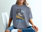 A blonde woman wears a grey t-shirt featuring a cartoon goat sliding down a mountain with the text It's All Downhill From Here.