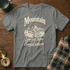 A gray t-shirt with a cream-colored graphic featuring mountains, pine trees, and text reading "Mountain Sunrise hike. Sunset arrival."