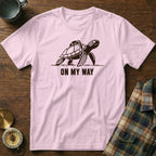 Pink t-shirt with a graphic of a turtle wearing a backpack and the text "ON MY WAY".