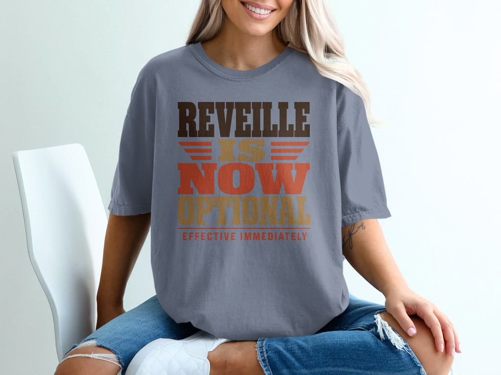 A woman wearing a gray t-shirt with brown, gold, and orange text that reads REVEILLE IS NOW OPTIONAL EFFECTIVE IMMEDIATELY.