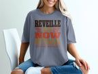 A woman wearing a gray t-shirt with brown, gold, and orange text that reads REVEILLE IS NOW OPTIONAL EFFECTIVE IMMEDIATELY.