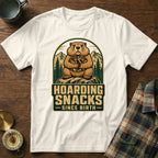 White t-shirt with a graphic of a groundhog hoarding nuts, with text "Hoarding Snacks Since Birth".
