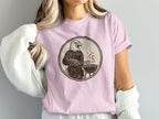 Eagle in apron grilling with tongs in circular graphic on pink t-shirt.