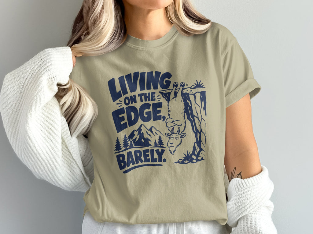 Khaki t-shirt with navy graphic of goat hanging from cliff, mountains, and text.