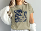 Khaki t-shirt with navy graphic of goat hanging from cliff, mountains, and text.