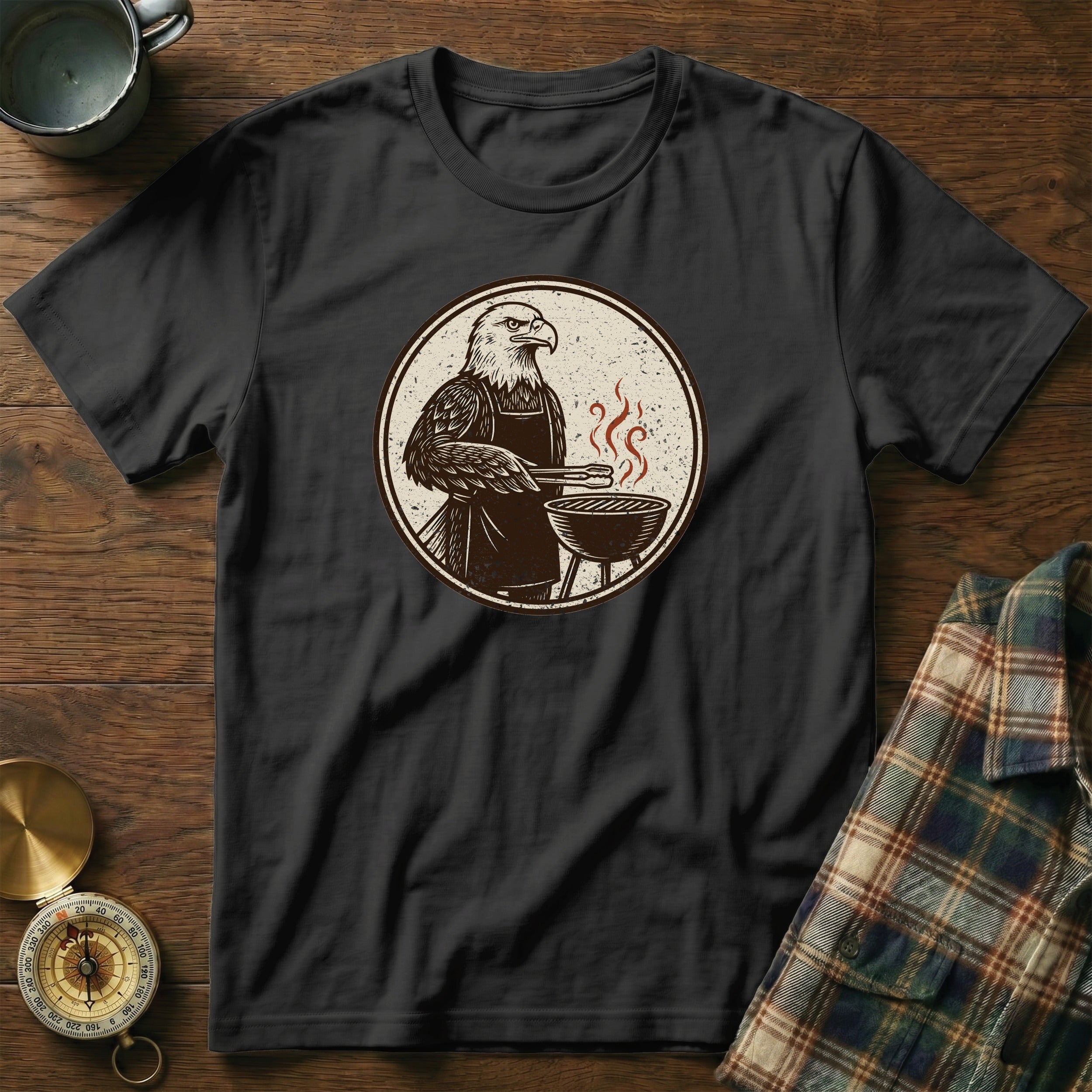Black t-shirt with eagle grilling on a circular graphic, laid on a wooden surface.