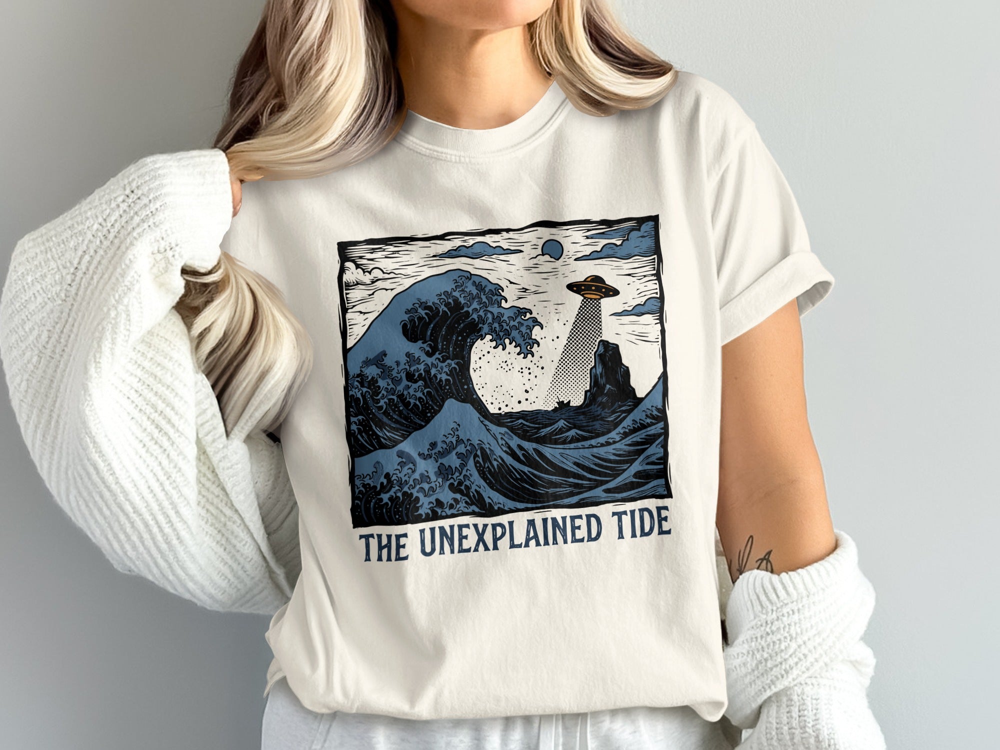 T-shirt with blue wave, UFO, lighthouse, and "The Unexplained Tide" text.