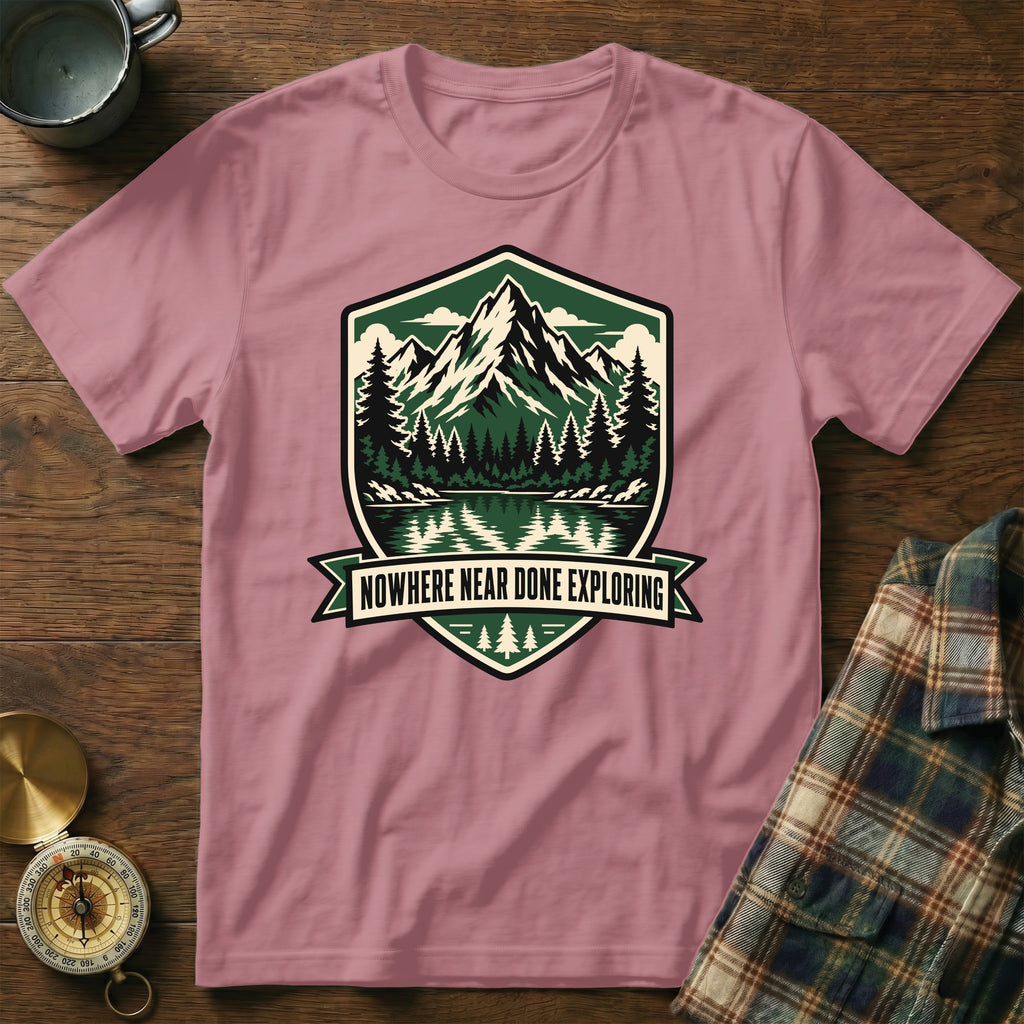Pink t-shirt with a shield logo featuring mountains, trees, and the text "Nowhere Near Done Exploring".