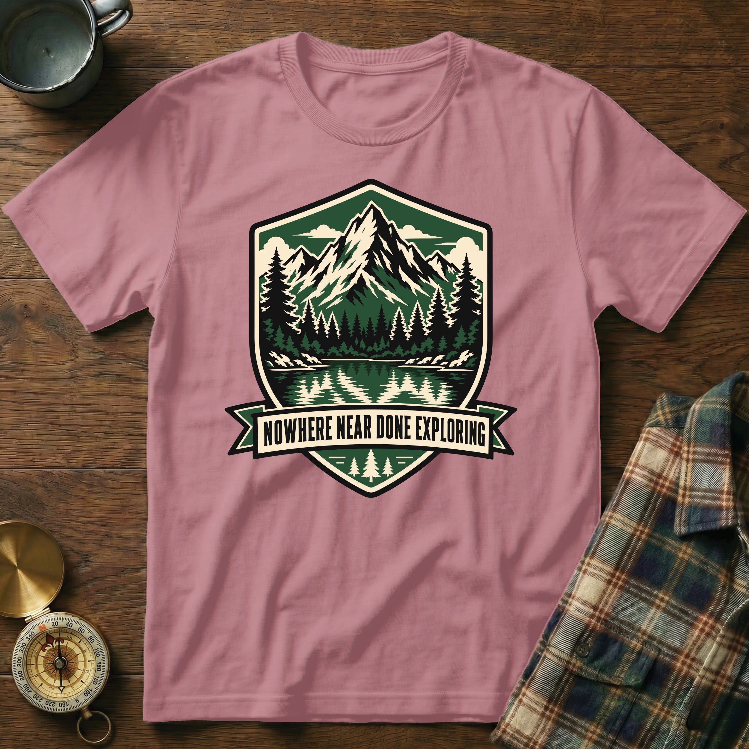 Pink t-shirt with a shield logo featuring mountains, trees, and the text "Nowhere Near Done Exploring".