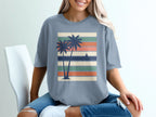 Blue t-shirt with graphic of two palm trees and sailboat on striped background.
