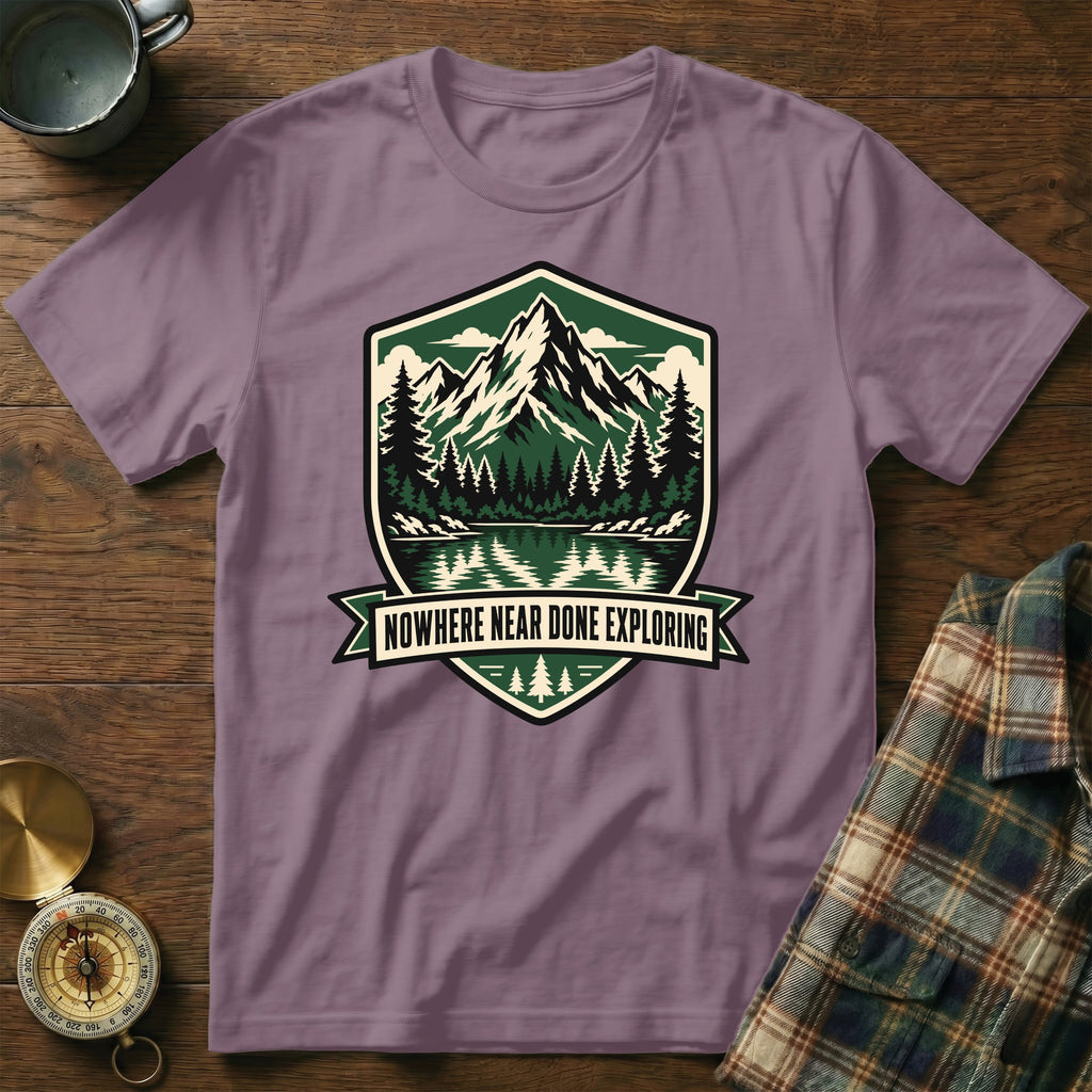 Purple t-shirt with a shield graphic of mountains, trees, and water, text "NOWHERE NEAR DONE EXPLORING".