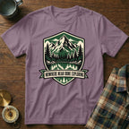 Purple t-shirt with a shield graphic of mountains, trees, and water, text "NOWHERE NEAR DONE EXPLORING".