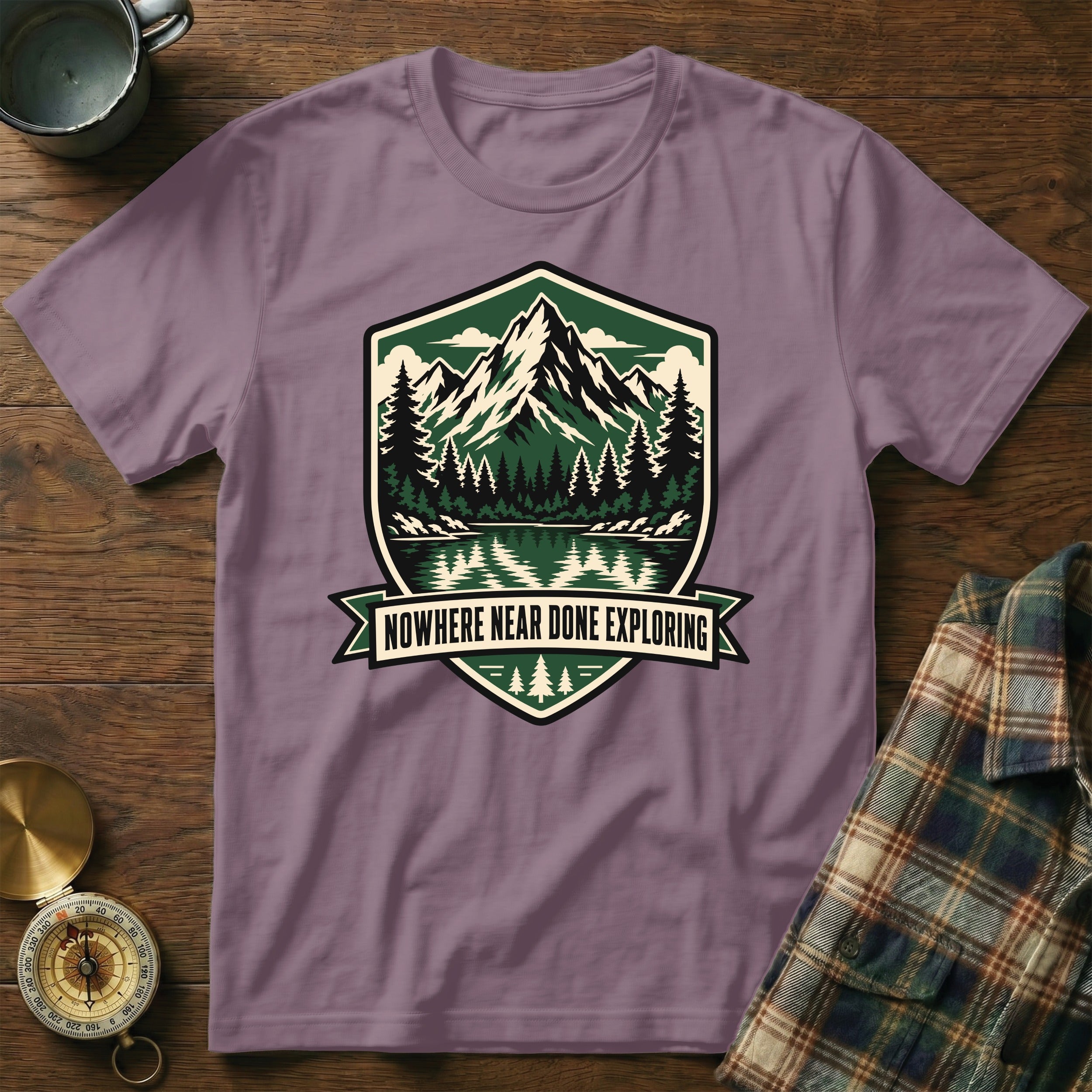 Purple t-shirt with a shield graphic of mountains, trees, and water, text "NOWHERE NEAR DONE EXPLORING".