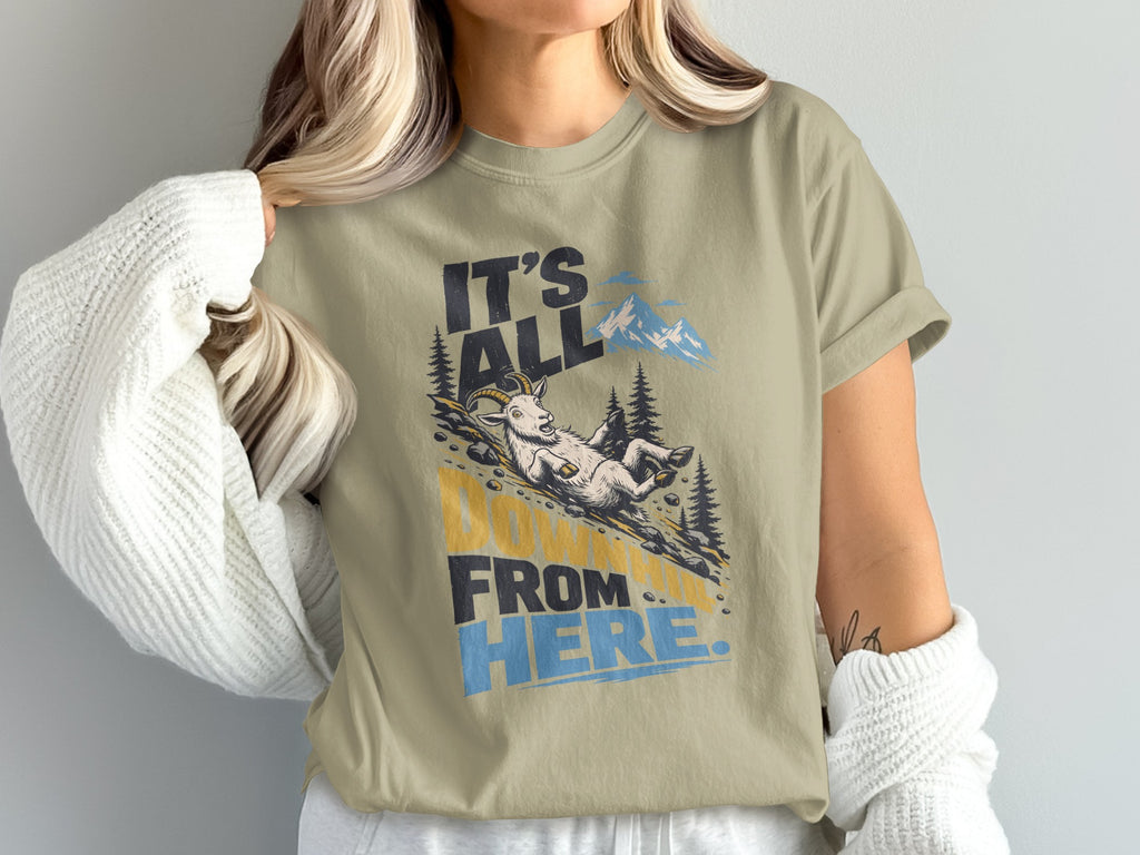 A person wearing an olive-green t-shirt printed with a graphic of a goat sliding downhill and the text It's All Downhill From Here.