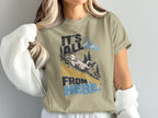 A person wearing an olive-green t-shirt printed with a graphic of a goat sliding downhill and the text It's All Downhill From Here.