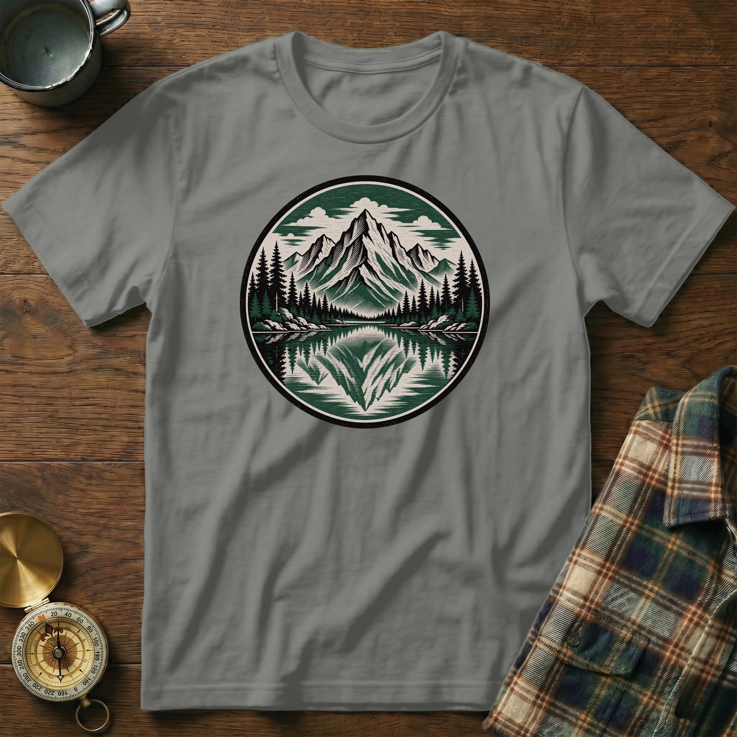 Grey t-shirt with a round mountain and forest graphic reflected in water.