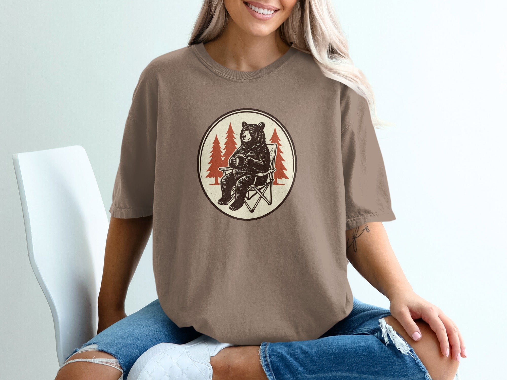 T-shirt with a bear in a camp chair holding a mug, surrounded by pine trees.