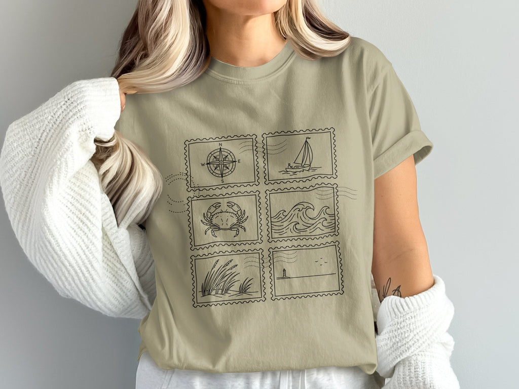 Light brown t-shirt with six postage stamp illustrations: compass, sailboat, crab, waves, grass, and lighthouse.