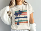 Cream t-shirt with retro palm trees and sailboat on striped background.