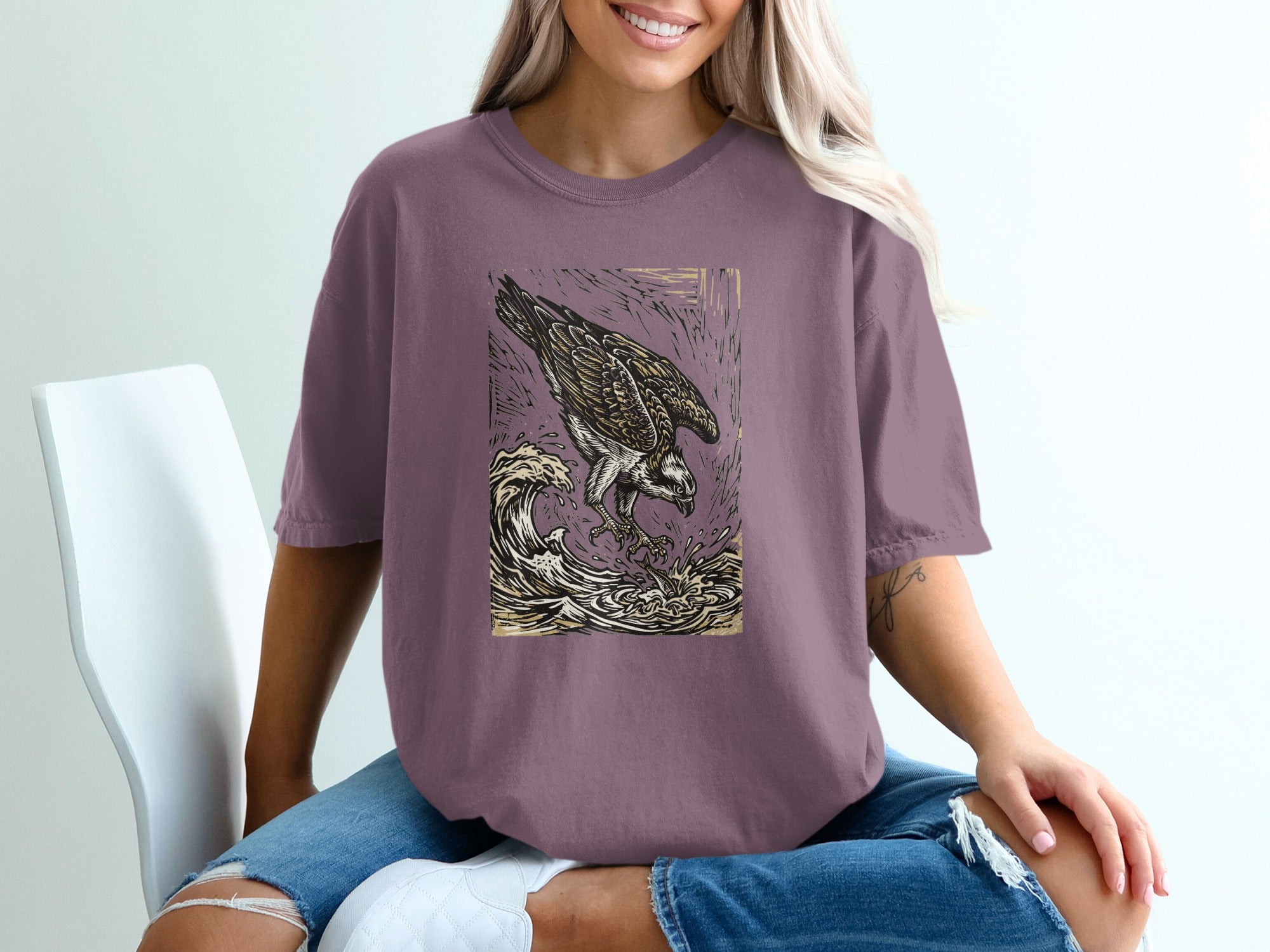 Osprey in flight over crashing waves, stylized graphic on a mauve t-shirt.