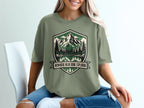 Olive green t-shirt with mountain, forest, and lake art and text "Nowhere Near Done Exploring."